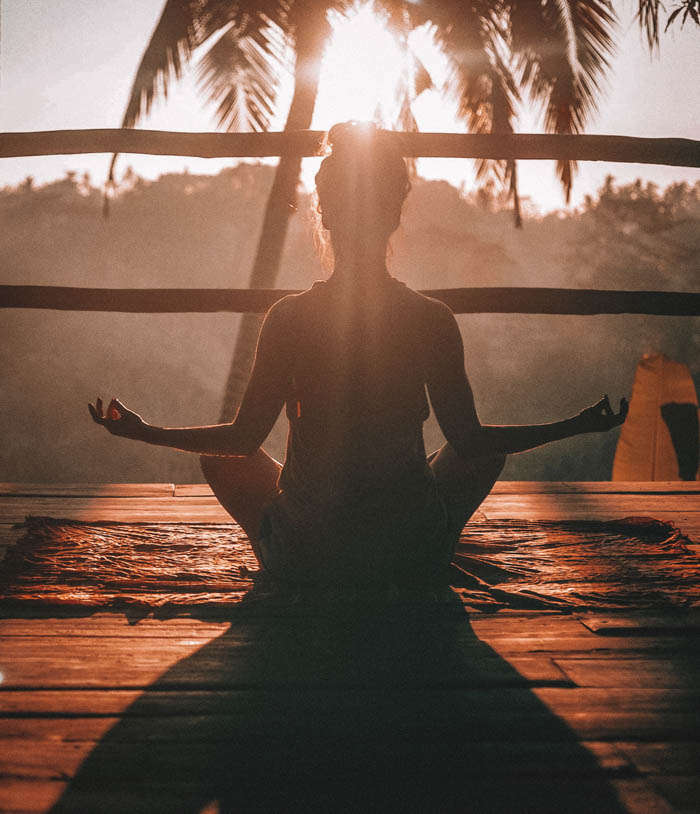A woman meditating at sunrise outside in front of a palm tree.