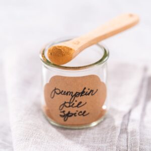 A jar with the words pumpkin pie spice written in black cursive lettering on a brown jar label, with a tiny wooden spoon holding a teaspoons worth of the homemade pumpkin pie seasoning.