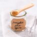 A jar with the words pumpkin pie spice written in black cursive lettering on a brown jar label, with a tiny wooden spoon holding a teaspoons worth of the homemade pumpkin pie seasoning.