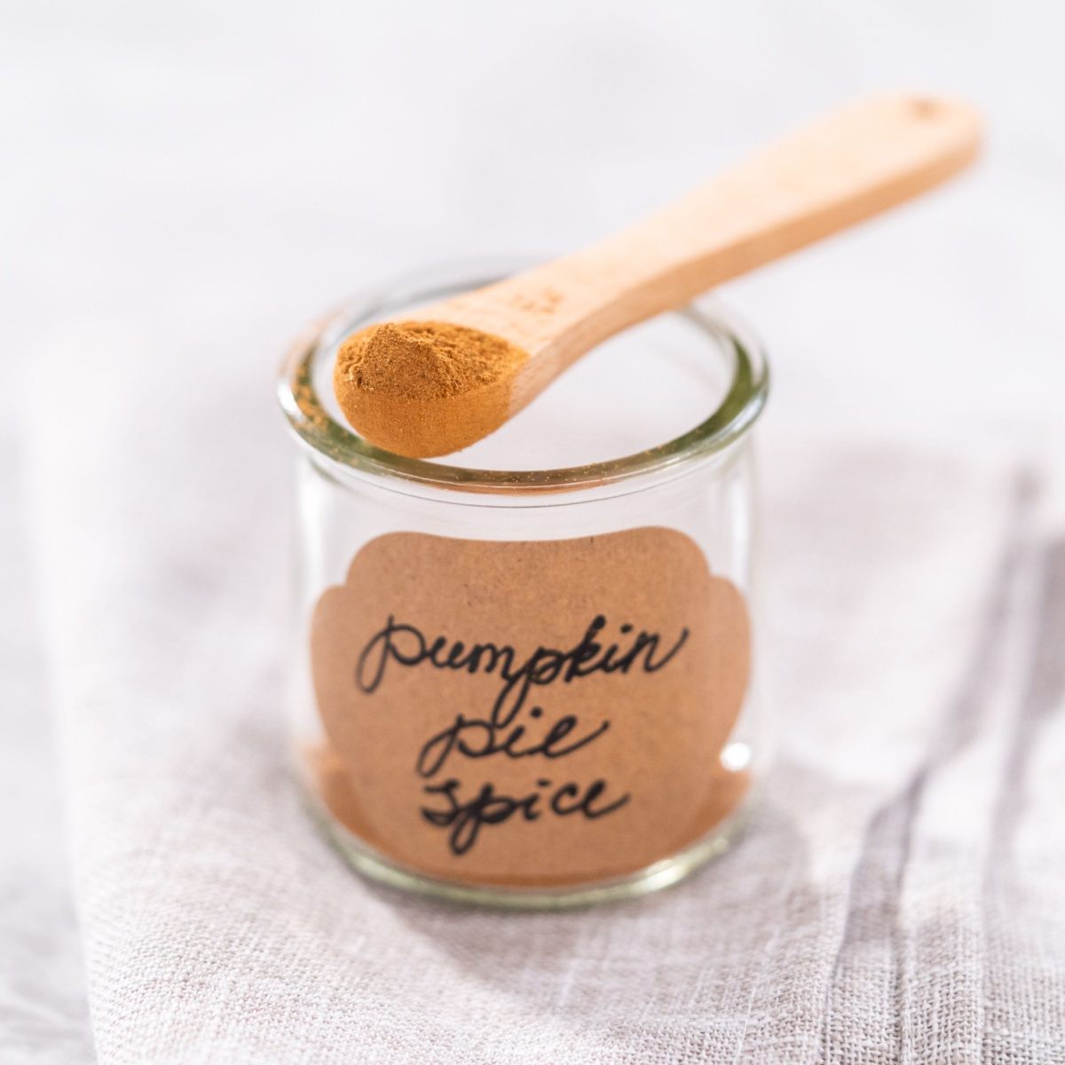 Homemade pumpkin pie seasoning jar A jar with the words pumpkin pie spice written in black cursive lettering on a brown jar label, with a tiny wooden spoon holding a teaspoons worth of the homemade pumpkin pie seasoning.